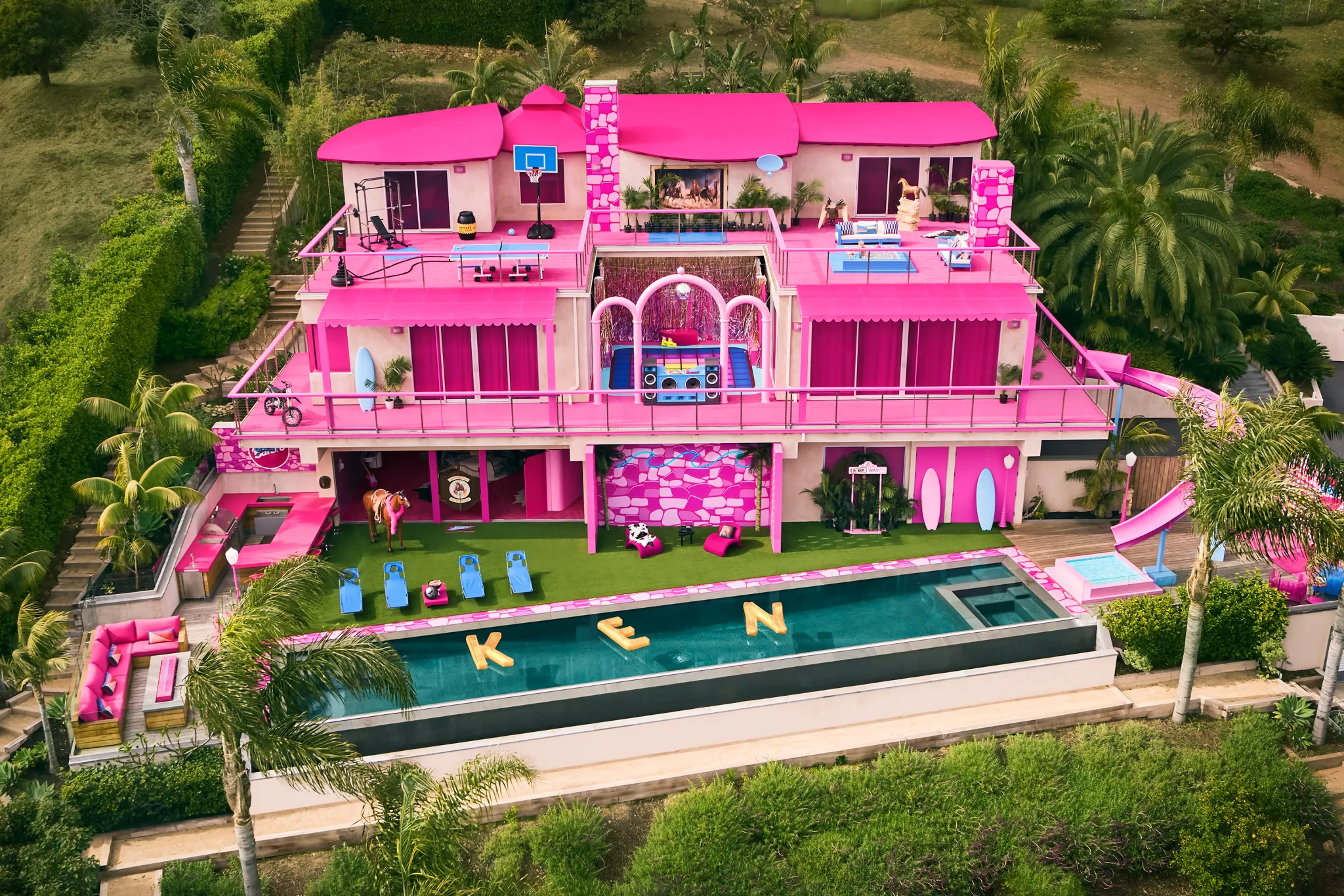 A real-life pink Malibu mansion styled as Barbie’s DreamHouse, featuring bright pink walls, pool floats spelling “KEN,” and palm trees surrounding the villa.