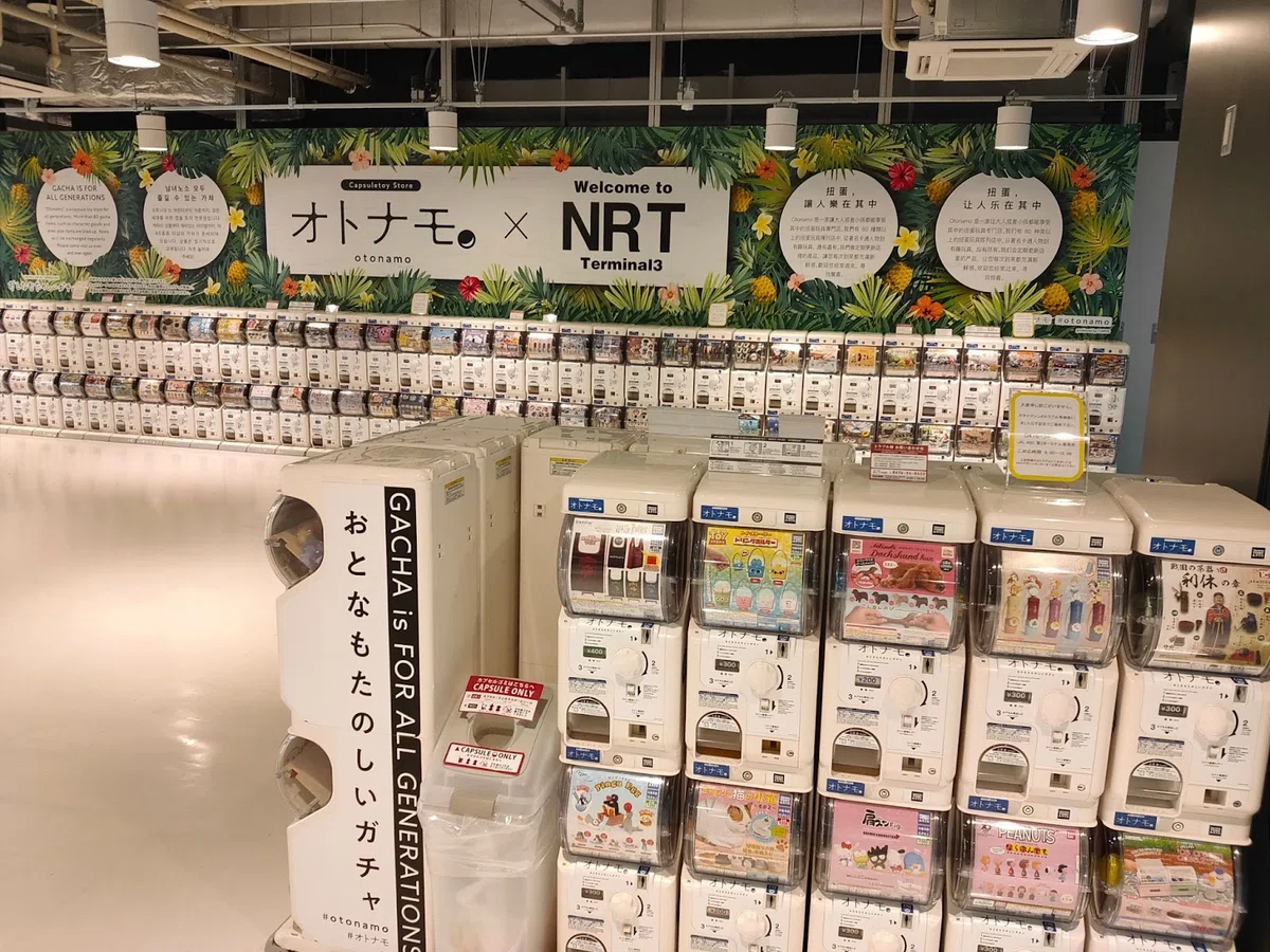Narita Airport Terminal 3 gacha zone with Otonamo × NRT wall graphics and long rows of capsule machines.