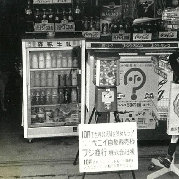 Vintage black-and-white storefront with a small capsule toy machine on a tripod next to a Pepsi cooler.